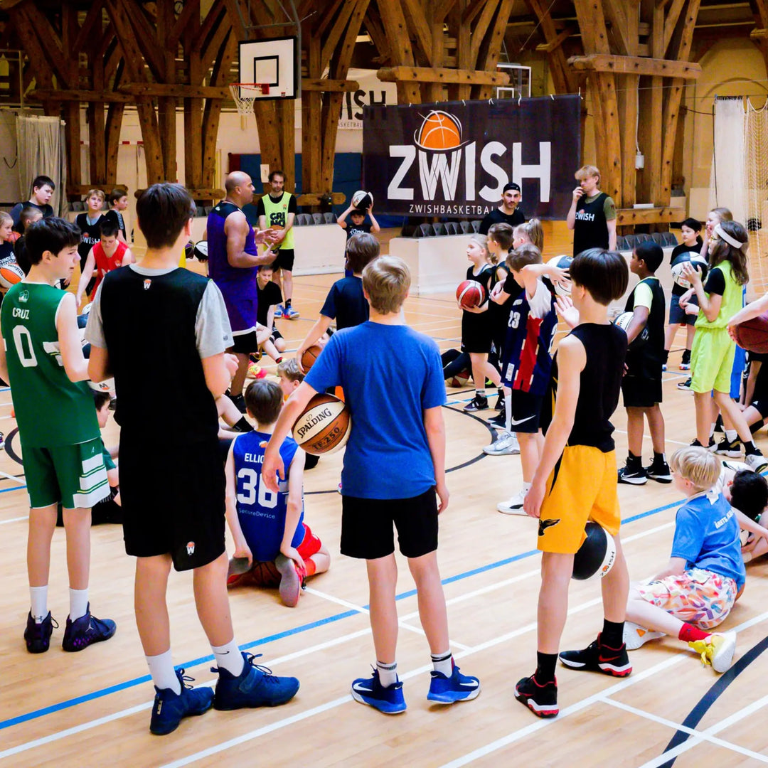 Young players in basketball jerseys gathered around a coach on court at a ZWISH basketball camp